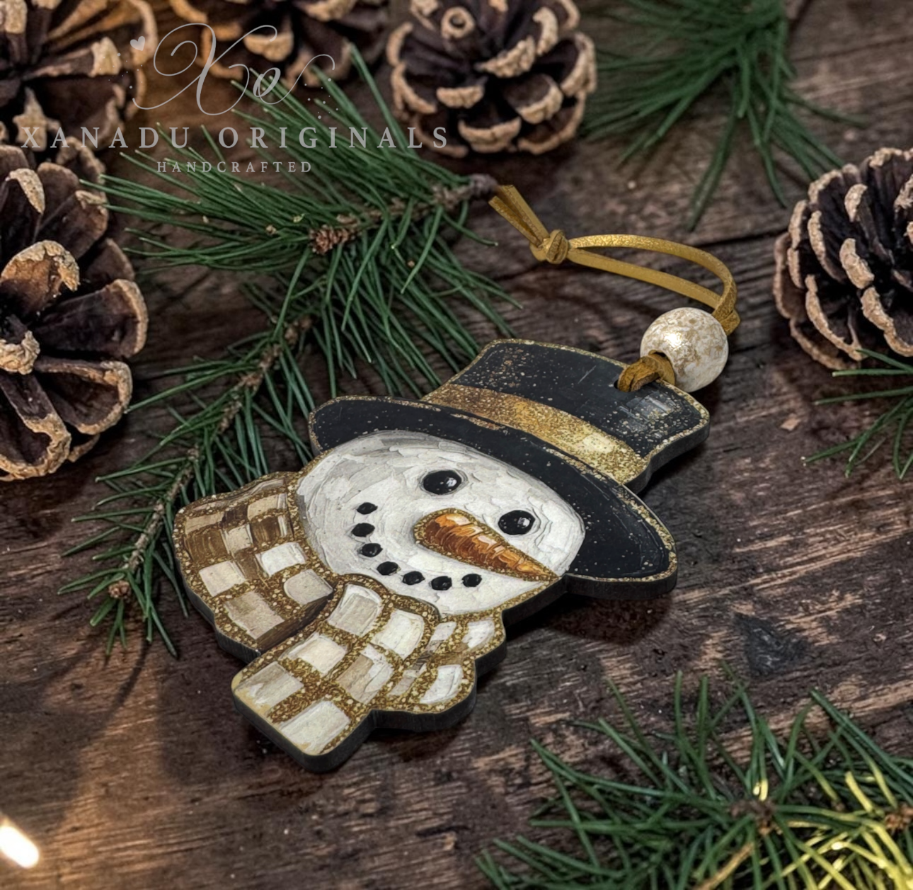 Handcrafted wooden snowman ornament with a black hat, white face and scarf, gold trim, and an orange carrot nose, hanging on a rustic wooden surface with pine branches and pinecones. Kanadu Originals watermark is visible.