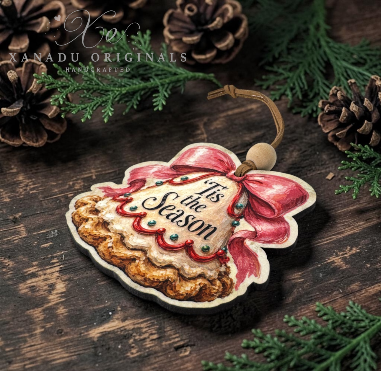 A Christmas ornament shaped like a pie with a red bow and the words 'It's the Season' on the front, displayed on a dark wooden surface with pinecones and evergreen branches. A watermark for 'Xanadu Originals Handcrafted' is visible in the upper left.