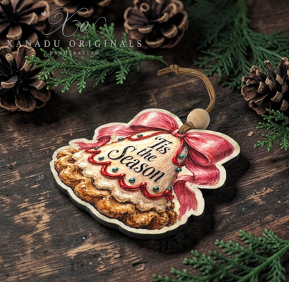 A Christmas ornament shaped like a pie with a red bow and the words 'It's the Season' on the front, displayed on a dark wooden surface with pinecones and evergreen branches. A watermark for 'Xanadu Originals Handcrafted' is visible in the upper left.