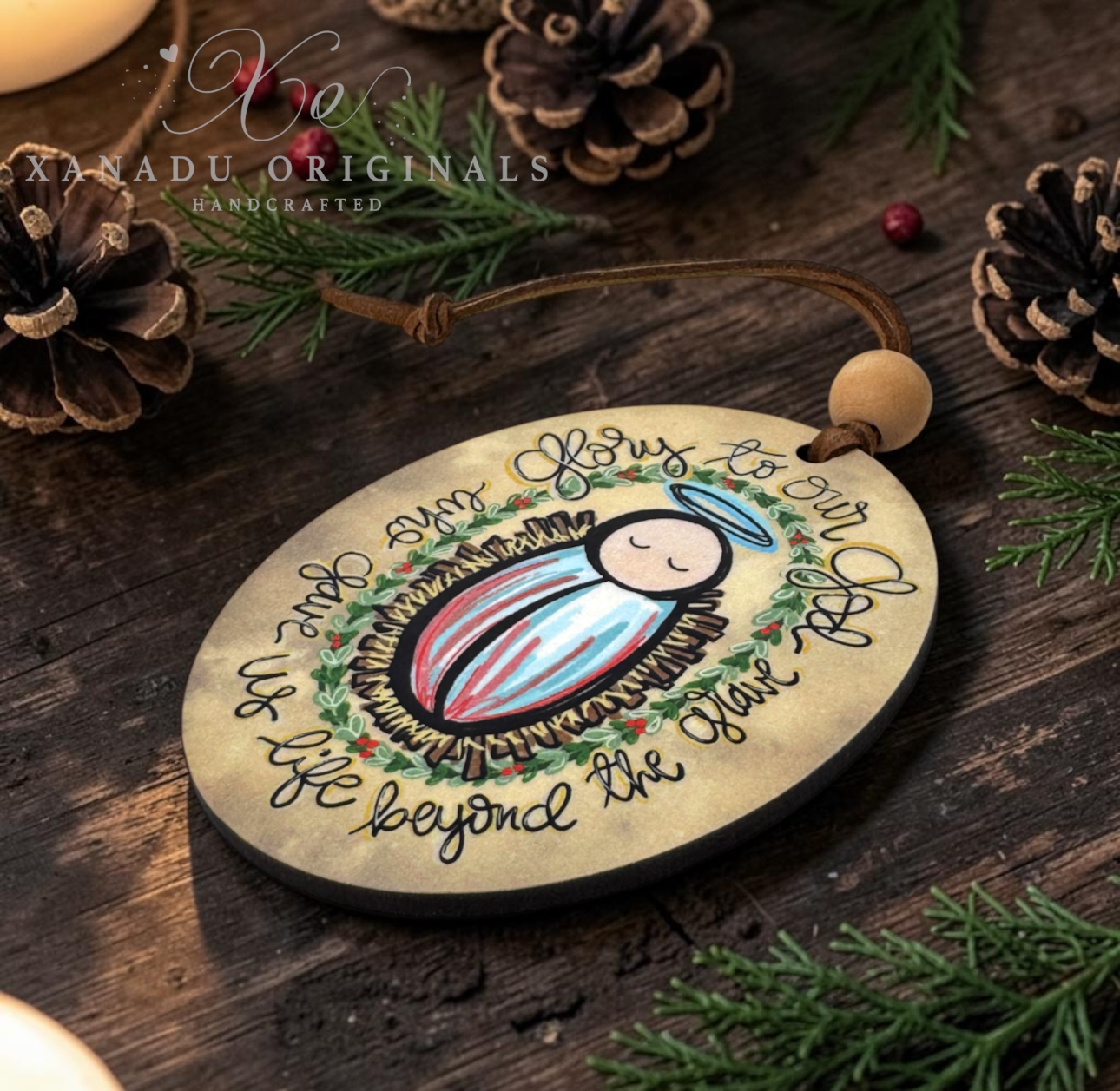 A round wooden Christmas ornament featuring a painted baby Jesus in a manger with handwritten text, hanging from a brown cord with a wooden bead. The ornament is styled on a dark wooden surface with pine branches, pinecones, and a warm candle glow, with the brand name Kanadu Originals visible in the upper left.