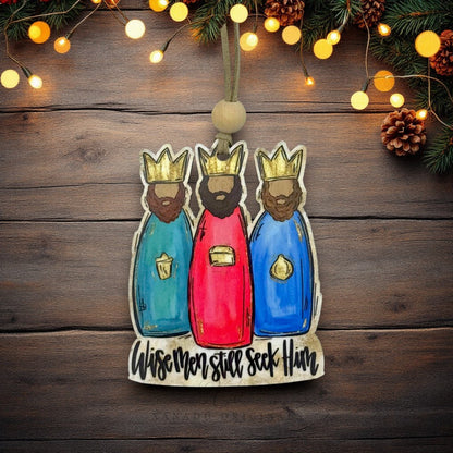 A hand-painted wooden Christmas ornament depicting the Three Wise Men, hanging against a rustic dark wood background decorated with pine branches, pinecones, and warm string lights.