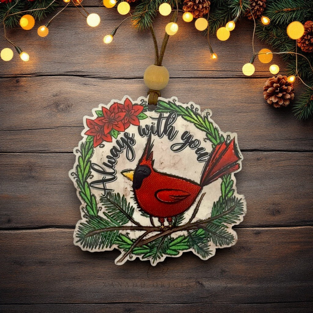 A wooden Christmas ornament featuring a red cardinal perched on a green pine branch, surrounded by the words 'Always with you' and red poinsettia flowers. The ornament hangs from a brown cord with a round wooden bead, set against a rustic dark wood background decorated with warm string lights and pinecones.