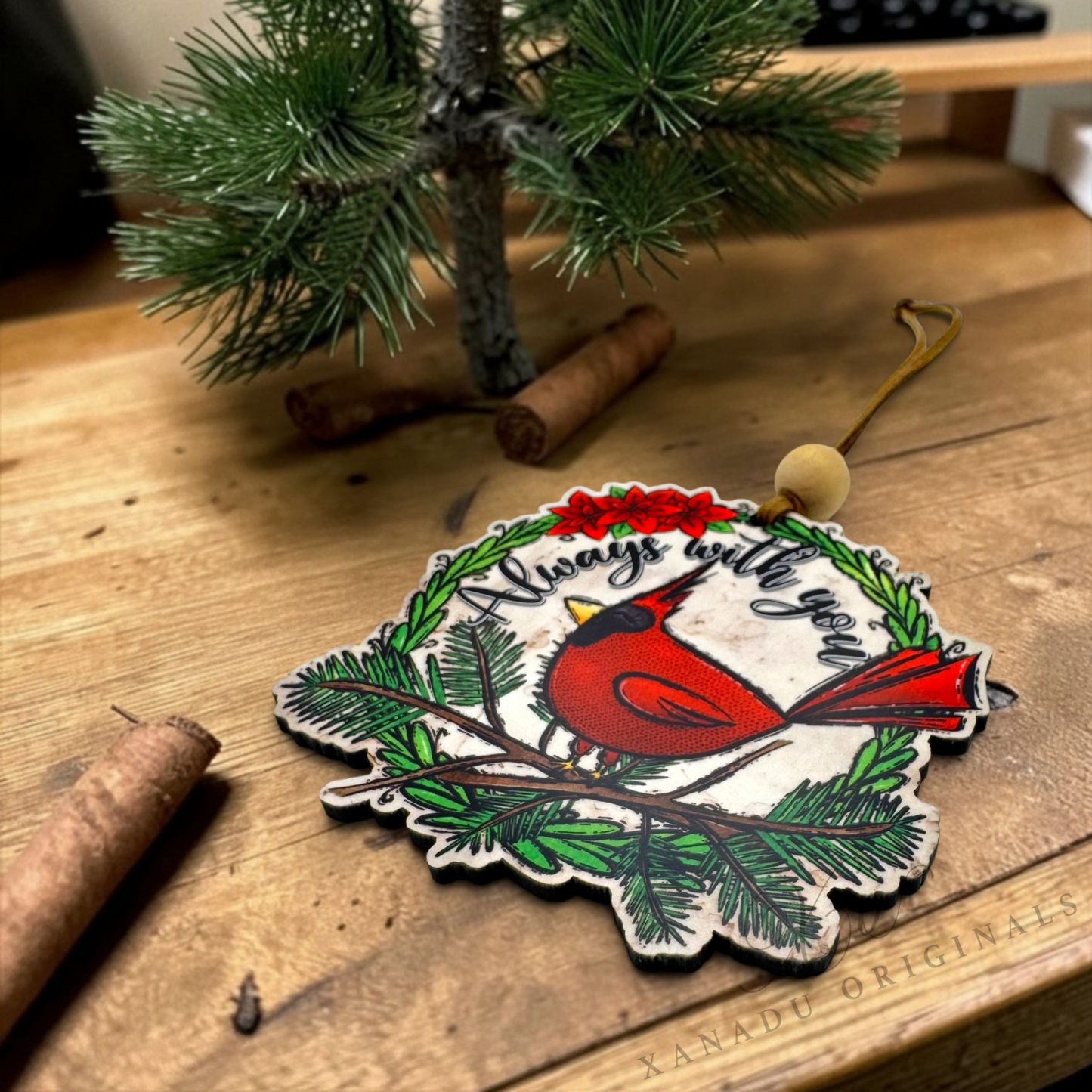 A laser-cut wooden Christmas ornament featuring a red cardinal on a green pine branch with the text 'Always with you' and red berries, displayed on a wooden tabletop next to a small pine tree and cinnamon sticks.