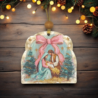 A Christmas ornament shaped like a rustic wooden sign with a painted nativity scene featuring Mary, Joseph, and baby Jesus under a large pink bow, hanging on a dark wood background with warm string lights and pine branches.