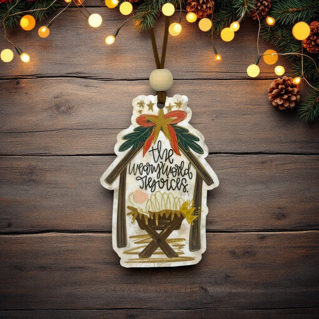 A hand-painted wooden Christmas nativity ornament hanging on a rustic dark wood background, decorated with a stable silhouette, gold stars, a red and gold bow, green holly leaves, and the words 'The weary world rejoices.' Christmas lights, pinecones, and evergreen branches decorate the top of the frame.