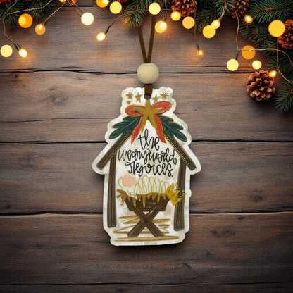 A hand-painted wooden Christmas nativity ornament hanging on a rustic dark wood background, decorated with a stable silhouette, gold stars, a red and gold bow, green holly leaves, and the words 'The weary world rejoices.' Christmas lights, pinecones, and evergreen branches decorate the top of the frame.