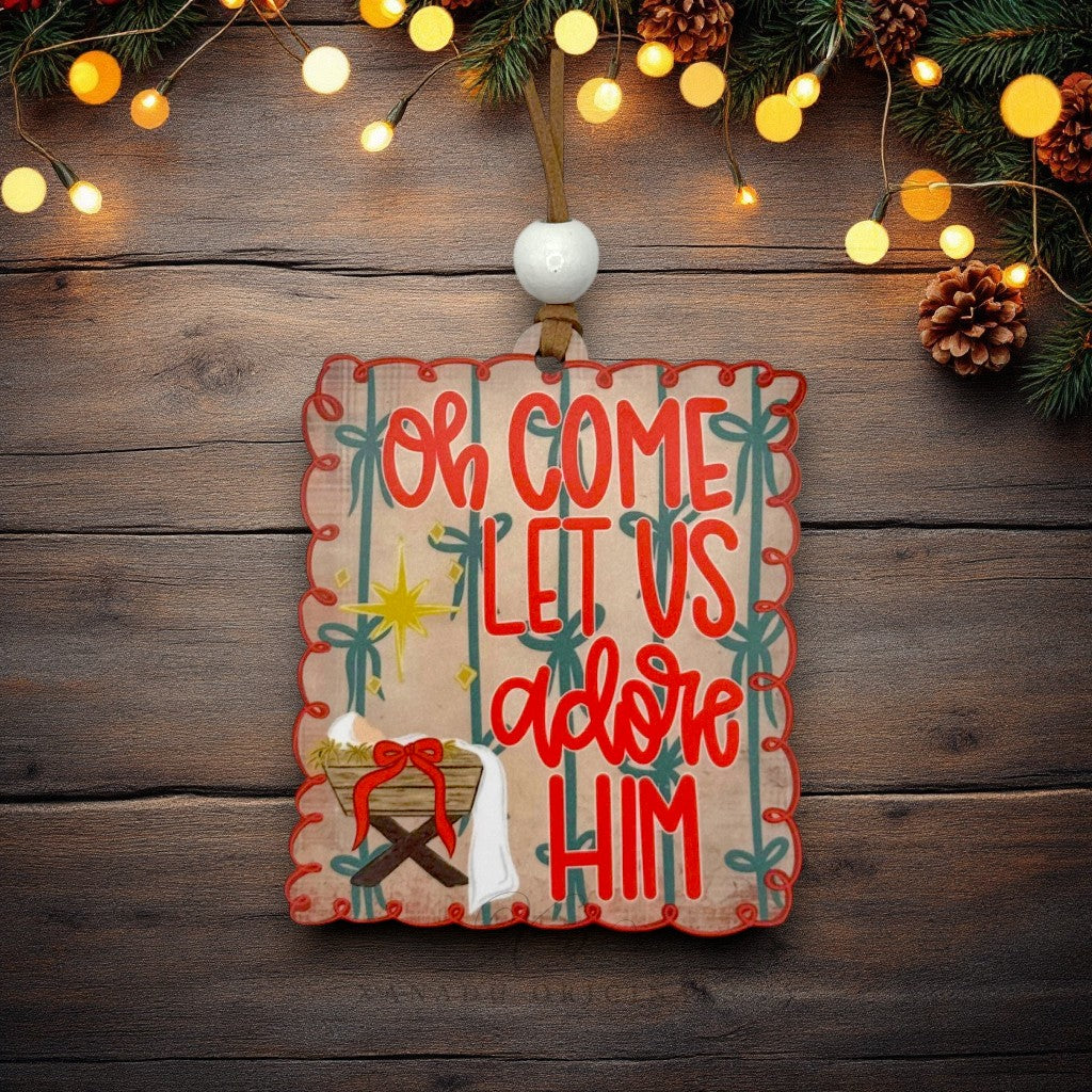 A rectangular wooden Christmas ornament with scalloped edges hangs on a rustic dark wood background. The ornament features the phrase 'Oh Come Let Us adore Him' in red lettering, decorated with red trim, green palm tree silhouettes, a gold star, and a small nativity scene at the bottom.