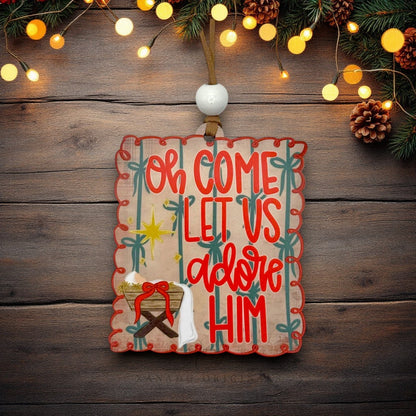 A rectangular wooden Christmas ornament with scalloped edges hangs on a rustic dark wood background. The ornament features the phrase 'Oh Come Let Us adore Him' in red lettering, decorated with red trim, green palm tree silhouettes, a gold star, and a small nativity scene at the bottom.