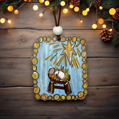 A rectangular wooden nativity ornament hanging on a rustic dark wood background, decorated with a hand-painted blue scene featuring a baby Jesus in a manger and a glowing gold star, surrounded by a border of gold and brown circles. The ornament is hung by a brown string with a white bead, and the background is decorated with pine branches, pinecones, and warm glowing fairy lights.