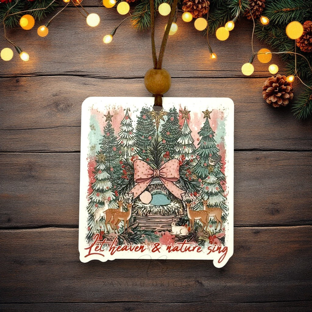 A rectangular Christmas ornament with rounded corners hangs from a brown string and wooden bead. The ornament features a festive illustration of a small nativity surrounded by decorated trees, deer, and the phrase 'Let heaven & nature sing' in red script.