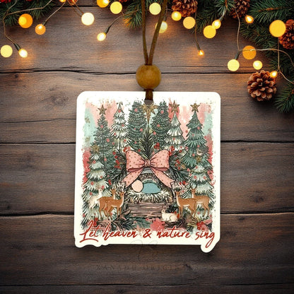 A rectangular Christmas ornament with rounded corners hangs from a brown string and wooden bead. The ornament features a festive illustration of a small nativity surrounded by decorated trees, deer, and the phrase 'Let heaven & nature sing' in red script.