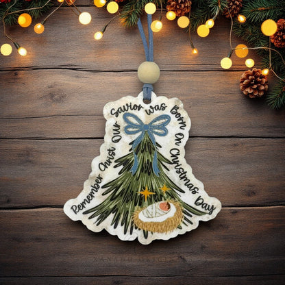 A decorative Christmas ornament shaped like a bell with a nativity tree illustration and handwritten text, hanging against a rustic wooden background with pine branches and warm string lights.