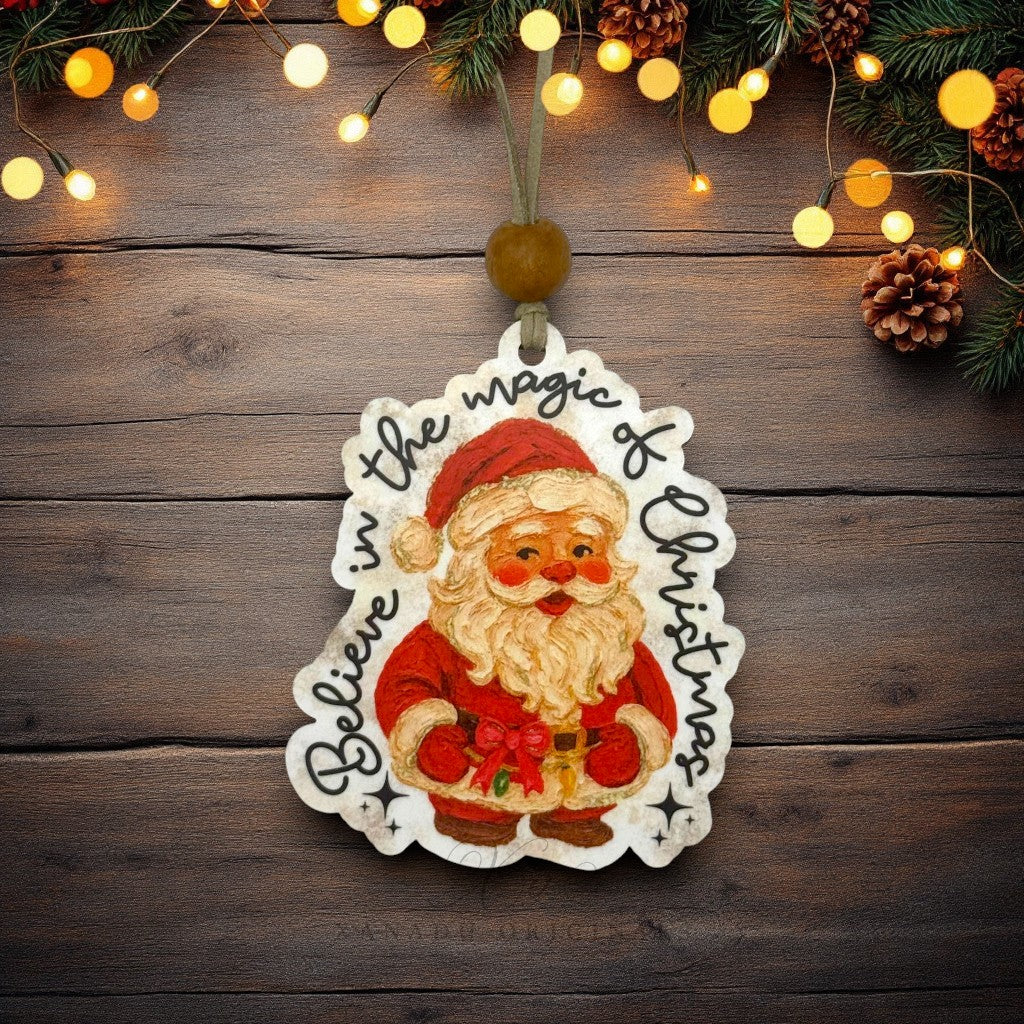 A die-cut Christmas ornament featuring an illustrated Santa Claus with the words 'Believe in the magic of Christmas' written around him, hanging against a rustic dark wood background with pine branches and warm string lights.