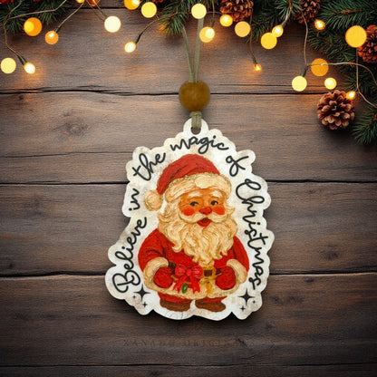 A die-cut Christmas ornament featuring an illustrated Santa Claus with the words 'Believe in the magic of Christmas' written around him, hanging against a rustic dark wood background with pine branches and warm string lights.