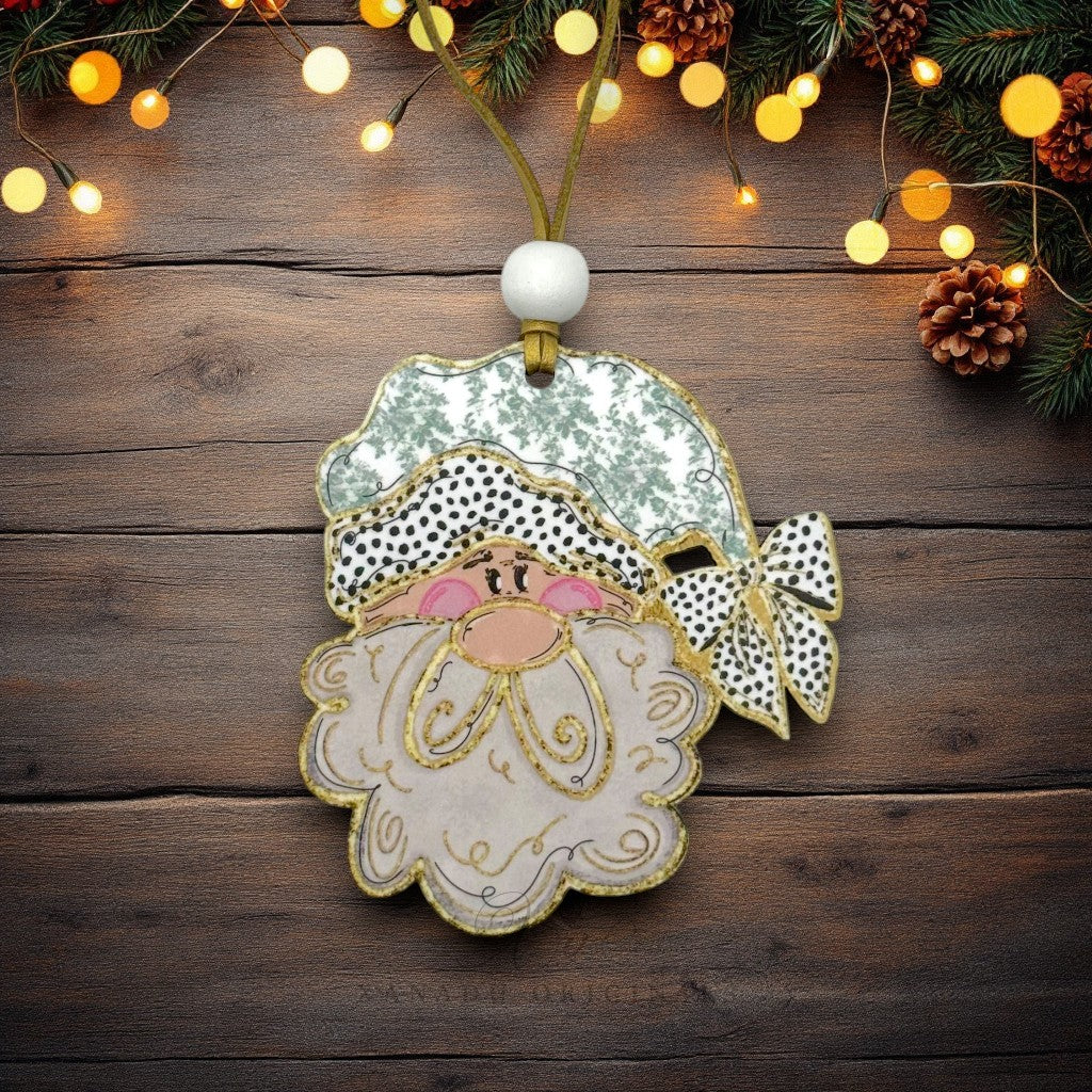 A handmade Christmas ornament shaped like Santa's face, featuring a white and green patterned hat, black-and-white polka dot bow, and fluffy white beard with gold glitter outlines, hanging on a rustic dark wood background with festive lights and pinecones.