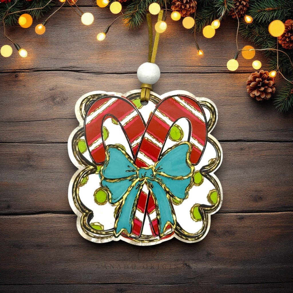 A Christmas ornament shaped like a gift box with two red-and-white striped candy canes, a turquoise bow, and gold trim, hanging on a dark wood surface with warm string lights and pine garland around the edges.