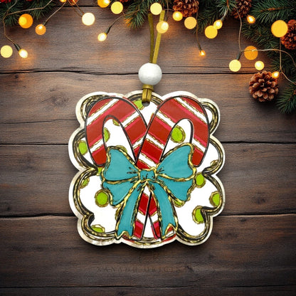 A Christmas ornament shaped like a gift box with two red-and-white striped candy canes, a turquoise bow, and gold trim, hanging on a dark wood surface with warm string lights and pine garland around the edges.