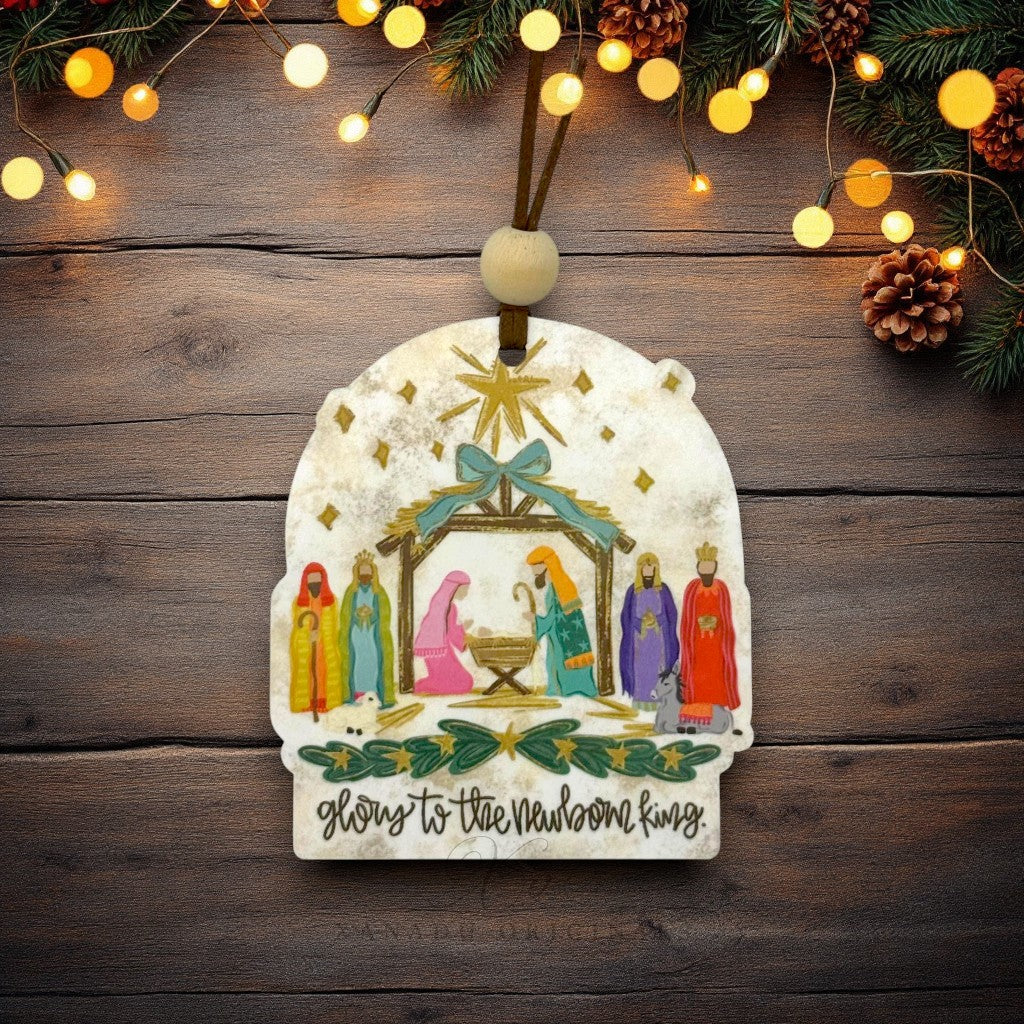 A Christmas ornament featuring a nativity scene with Mary, Joseph, baby Jesus, and three wise men, hanging on a rustic dark wood background with warm string lights and pine branches.