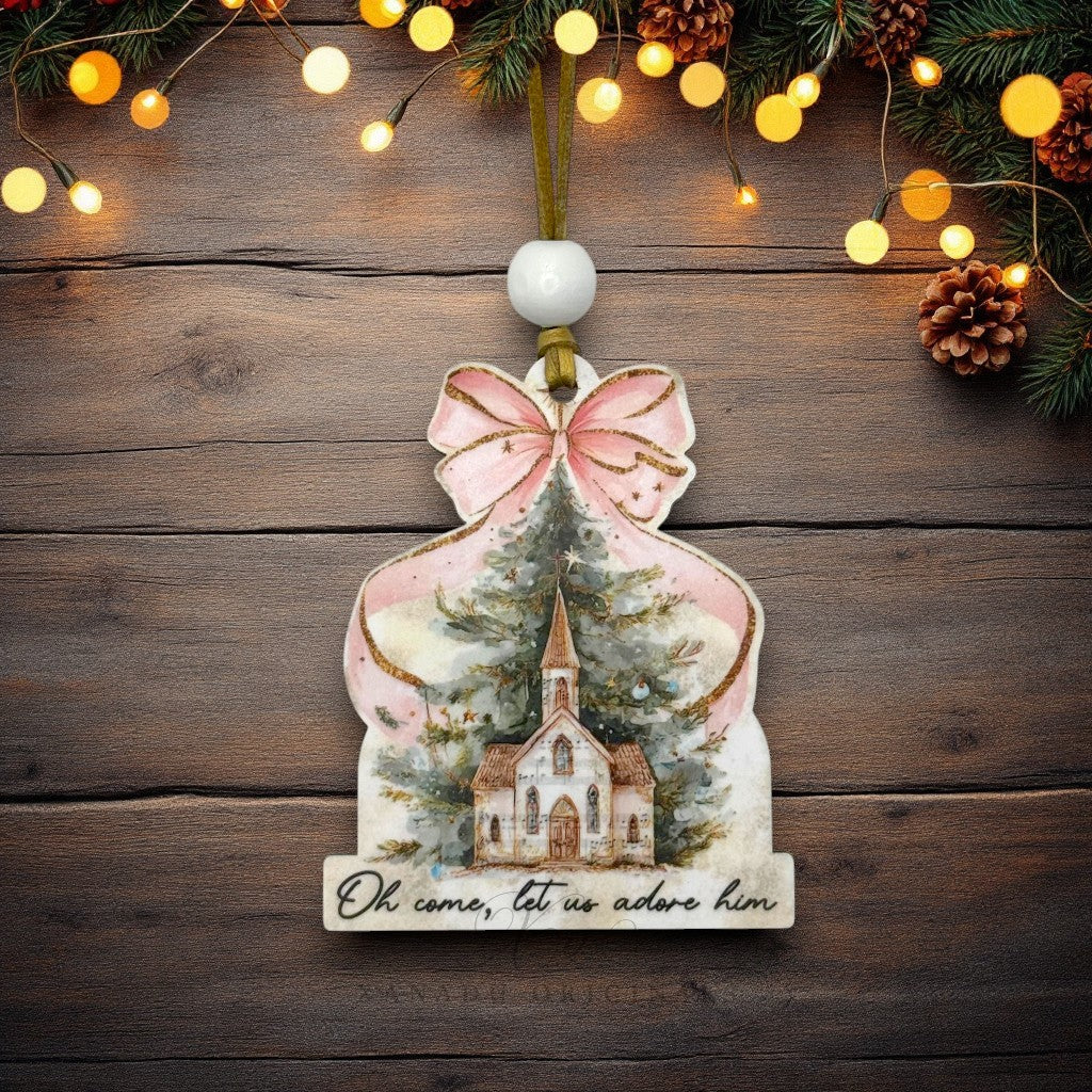 A Christmas ornament shaped like a gift box with a pink bow, featuring an illustration of a church and Christmas tree, hanging on a rustic wooden background with warm string lights and pinecones.