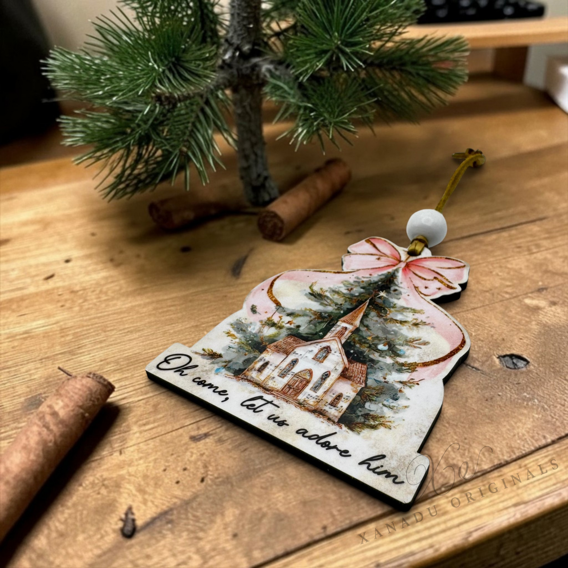 A Christmas ornament shaped like a house with a church illustration and the text 'Oh, how, let us adore him,' displayed on a wooden tabletop with cinnamon sticks and a small evergreen branch nearby.