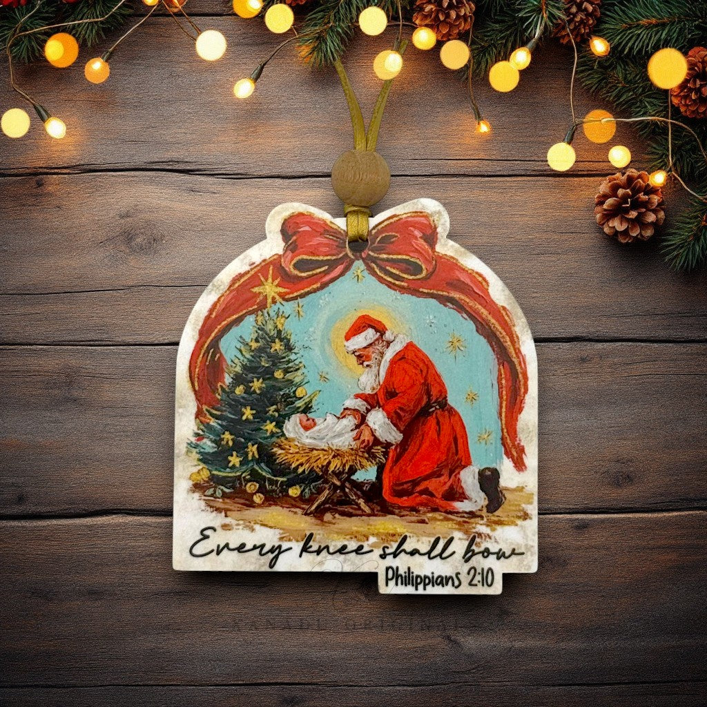 A personalized Christmas ornament featuring an illustration of Santa Claus kneeling by a baby Jesus next to a decorated Christmas tree, with the text 'Every knee shall bow' and 'Philippians 2:10'. The ornament is hung on a dark wood background with festive lights, pinecones, and evergreen branches.