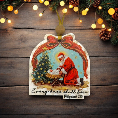 A personalized Christmas ornament featuring an illustration of Santa Claus kneeling by a baby Jesus next to a decorated Christmas tree, with the text 'Every knee shall bow' and 'Philippians 2:10'. The ornament is hung on a dark wood background with festive lights, pinecones, and evergreen branches.