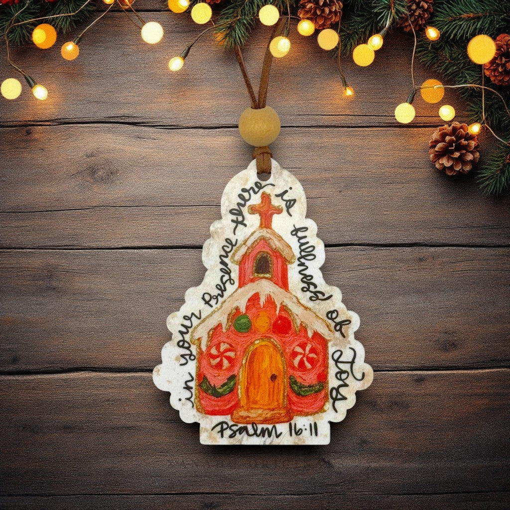 A hand-painted wooden Christmas ornament shaped like a church, decorated with colorful icing-style details and the Bible verse Psalm 16:11, hanging against a rustic dark wood background with warm string lights and pine branches at the top.