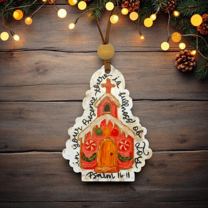 A hand-painted wooden Christmas ornament shaped like a church, decorated with colorful icing-style details and the Bible verse Psalm 16:11, hanging against a rustic dark wood background with warm string lights and pine branches at the top.