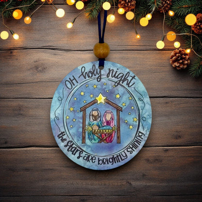 A round wooden Christmas ornament featuring a painted nativity scene with the text 'Oh holy night the stars are brightly shining'. The ornament hangs from a dark blue ribbon and a wooden bead, displayed on a rustic wooden background decorated with pine branches and warm string lights.