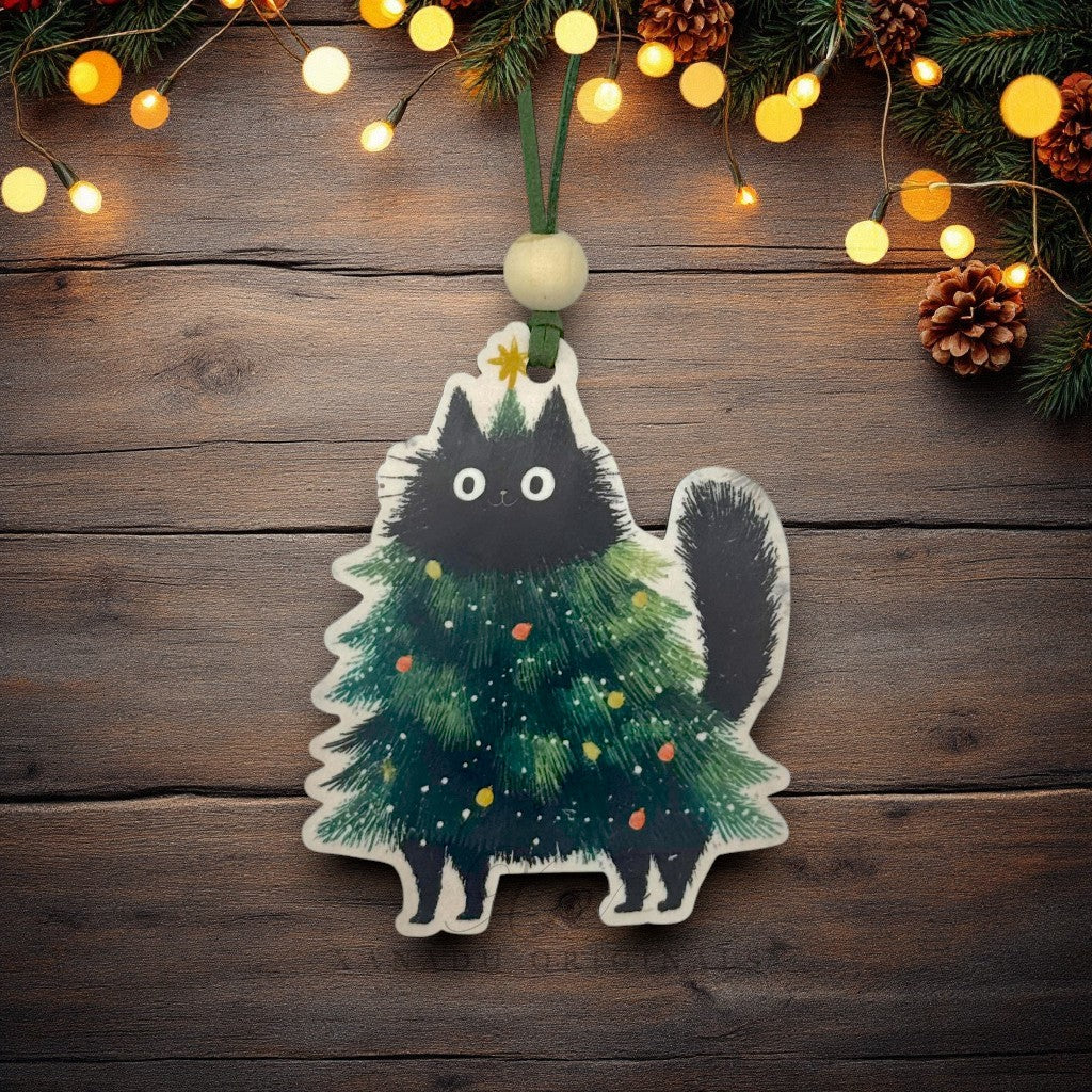 A Christmas ornament shaped like a black cat standing on a decorated green Christmas tree, hanging from a green cord with a cream bead on a rustic dark wood background with warm string lights and pine branches.