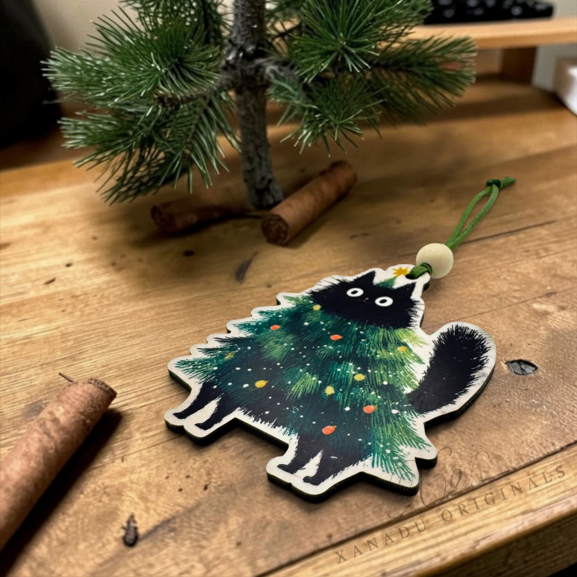 A wooden Christmas ornament shaped like a black cat dressed in a green Christmas tree with colorful ornaments, resting on a wooden tabletop next to cinnamon sticks and a small pine tree branch.