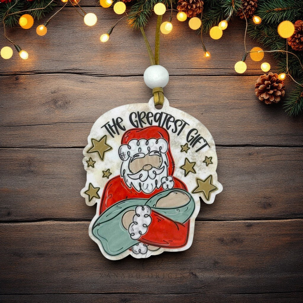 A hand-painted wooden Christmas ornament featuring a whimsical Santa Claus holding baby Jesus, with the text 'THE GREATEST GIFT' and gold stars. The ornament is hung on a dark wood background decorated with pine branches, pinecones, and warm glowing fairy lights.