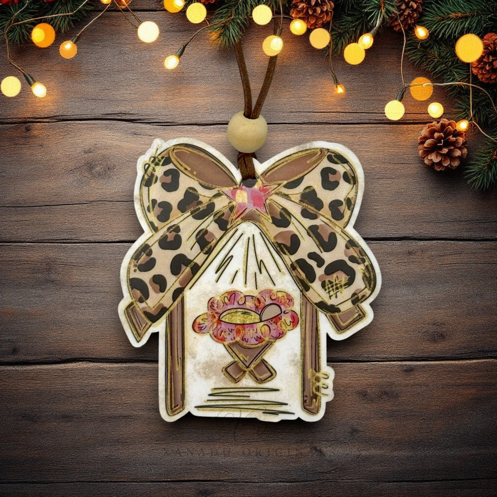 A decorative manger-shaped Christmas ornament with baby Jesus with a leopard-print blanket, a pink star, and a cheetah bow, hanging on a rustic dark wood background with warm string lights and pine branches.
