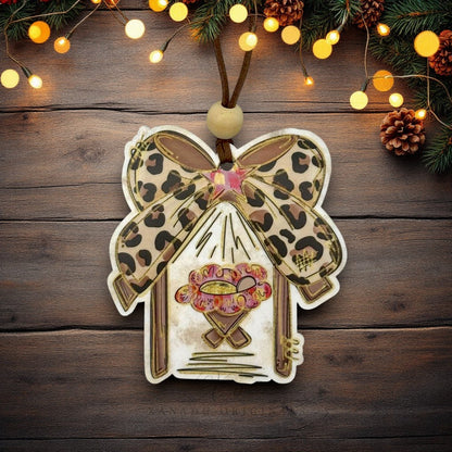 A decorative manger-shaped Christmas ornament with baby Jesus with a leopard-print blanket, a pink star, and a cheetah bow, hanging on a rustic dark wood background with warm string lights and pine branches.