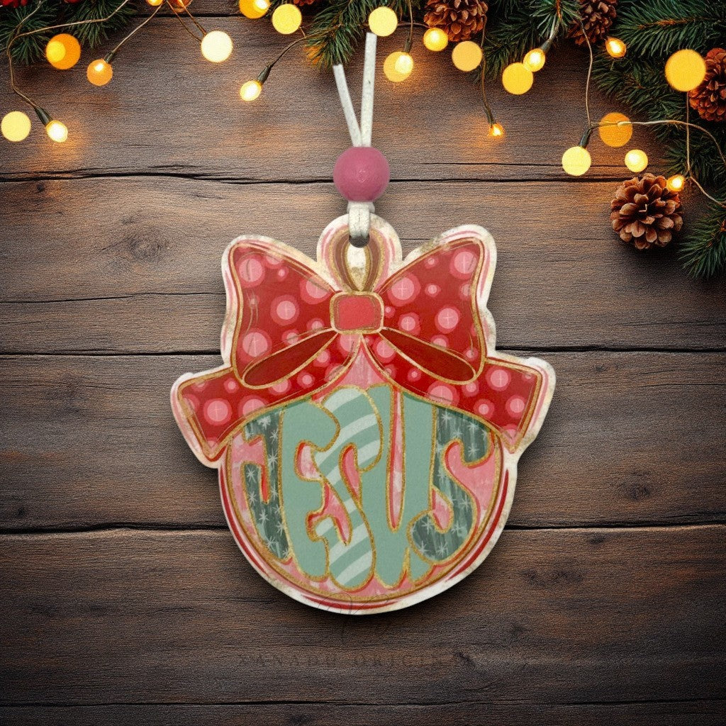 A personalized Christmas ornament shaped like a bell with a bow, hanging against a rustic dark wood background decorated with pine branches, pinecones, and warm string lights. The ornament features the name 'JESUS' in stylized green and white lettering on a pink background, with a red polka-dot bow on top.