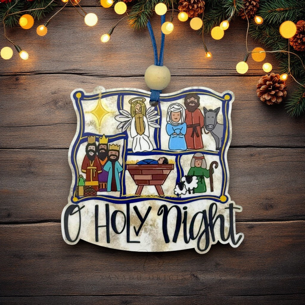 A decorative wooden Christmas ornament shaped like a scroll with a nativity scene and the words “O Holy Night” in script, hanging against a rustic wooden background with string lights and pine branches.