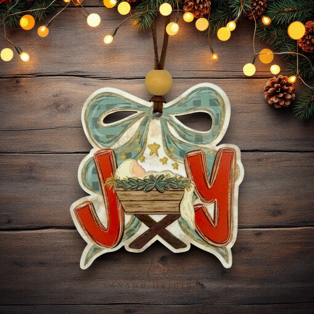 A decorative Christmas ornament shaped like the letter J and Y with a nativity scene and bow, hanging on a rustic dark wood background with pine branches and warm string lights.