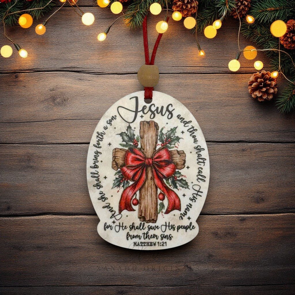 An oval Christmas ornament with a printed wooden cross, red bow, holly leaves, and Bible verse text, hanging on a rustic wood surface with warm string lights and pine branches around it.