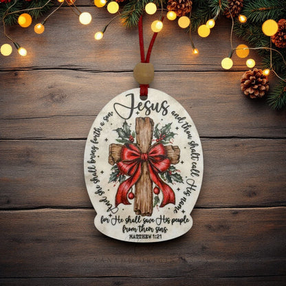 An oval Christmas ornament with a printed wooden cross, red bow, holly leaves, and Bible verse text, hanging on a rustic wood surface with warm string lights and pine branches around it.