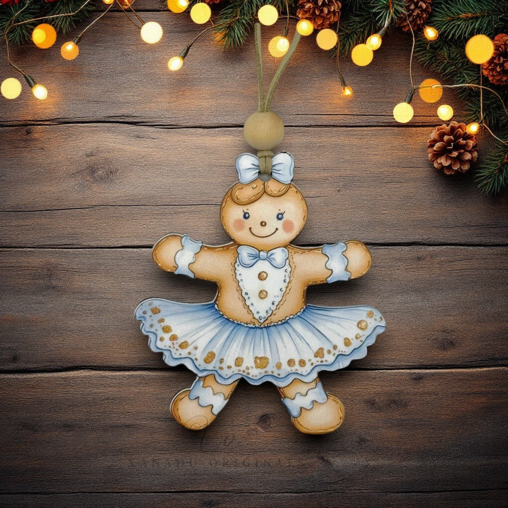 A hand-painted gingerbread girl Christmas ornament in a ballerina dress, hanging against a rustic dark wood background decorated with pine branches and warm string lights.