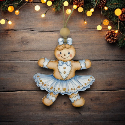A hand-painted gingerbread girl Christmas ornament in a ballerina dress, hanging against a rustic dark wood background decorated with pine branches and warm string lights.