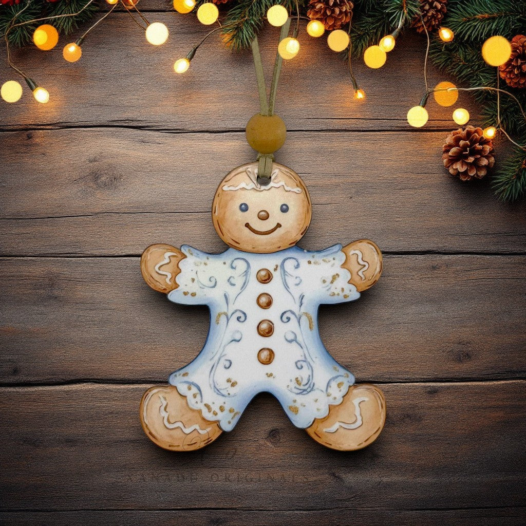 A hand-painted gingerbread man Christmas ornament hangs on a rustic dark wood background, decorated with light blue icing, gold buttons, and white icing details. The scene is decorated with pine branches and warm glowing fairy lights.