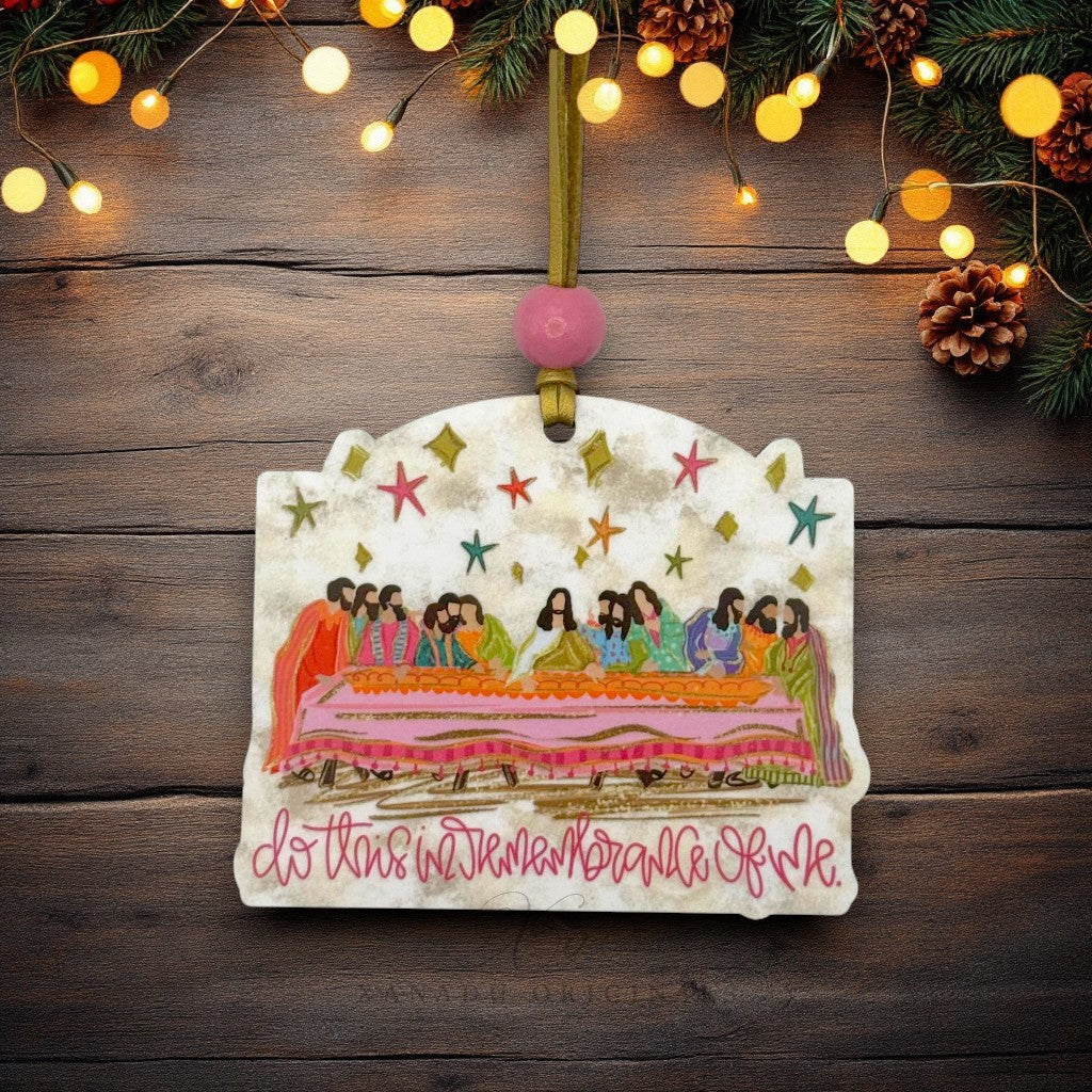 A hand-painted ceramic Christmas ornament featuring the Last Supper scene with the text 'do this in memory of me' in pink script. The ornament is hanging from a gold ribbon with a pink bead, set against a rustic dark wood background decorated with pine branches and warm string lights.