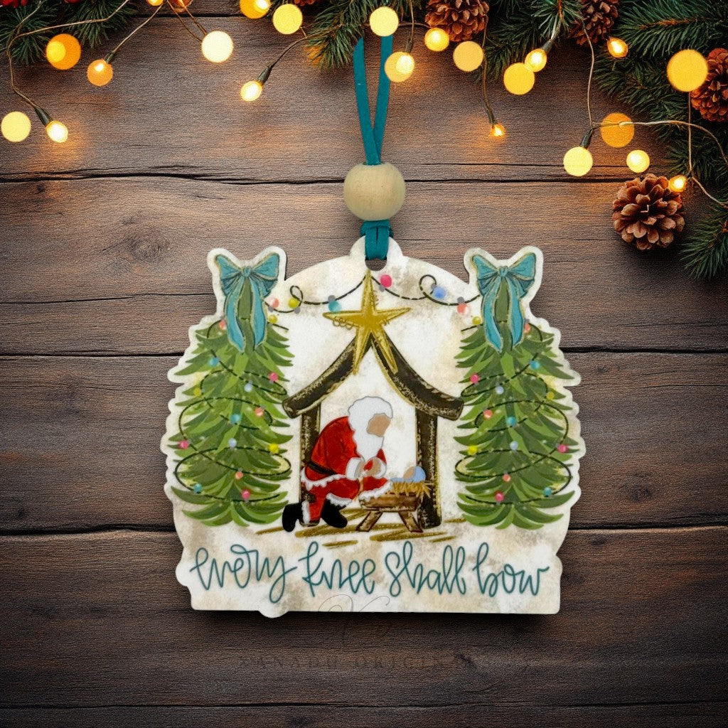A hand-painted ceramic Christmas ornament featuring Santa in a stable with two decorated Christmas trees and the text 'Every knee shall bow'. The ornament is hung on a dark wood background with festive lights and pinecones.