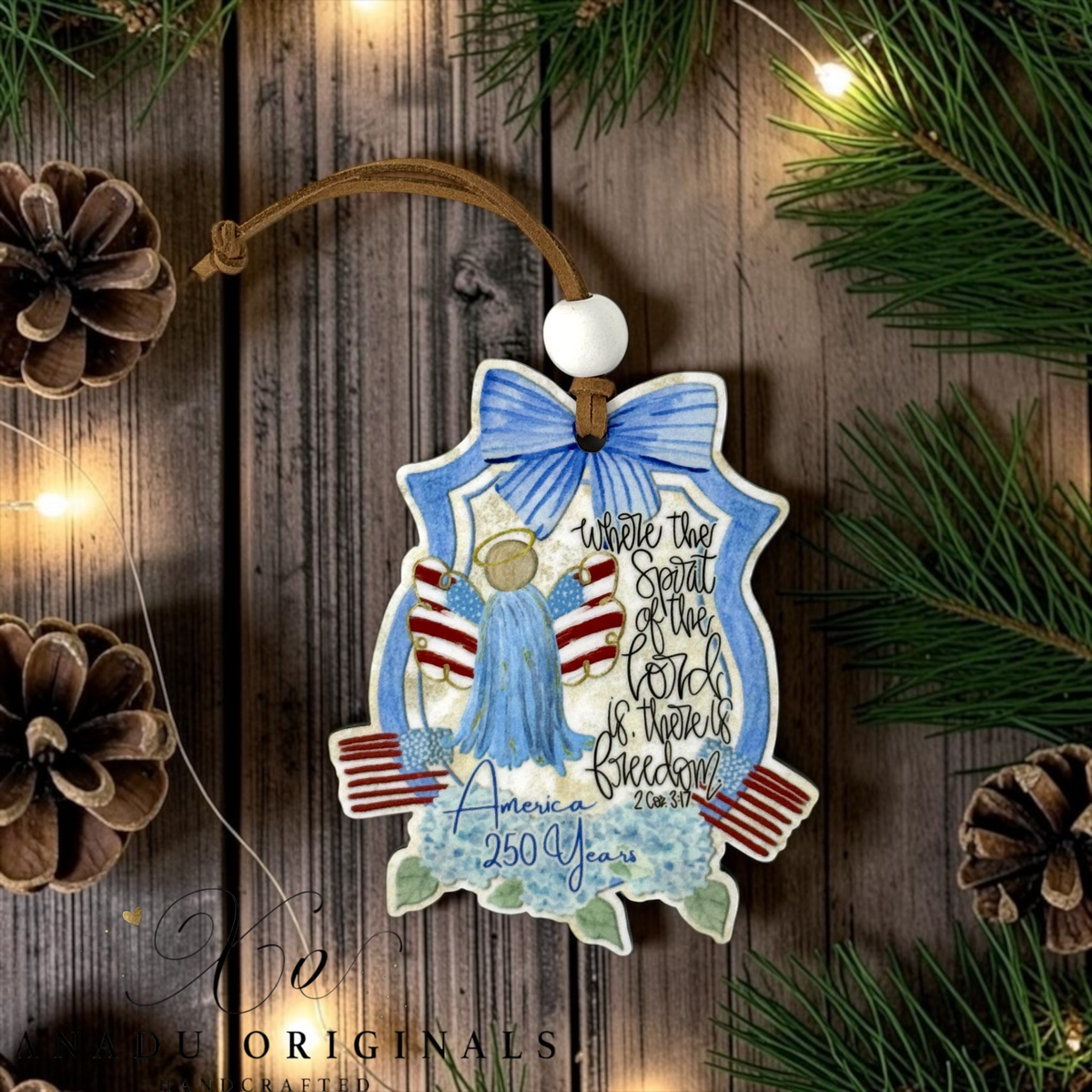 America 250 Years wood Christmas ornament featuring a blue angel with red and white striped wings, a blue bow, and patriotic elements, hanging on a rustic wooden background with pine branches and fairy lights. Where The Spirit of the Lord is there is freedom.
