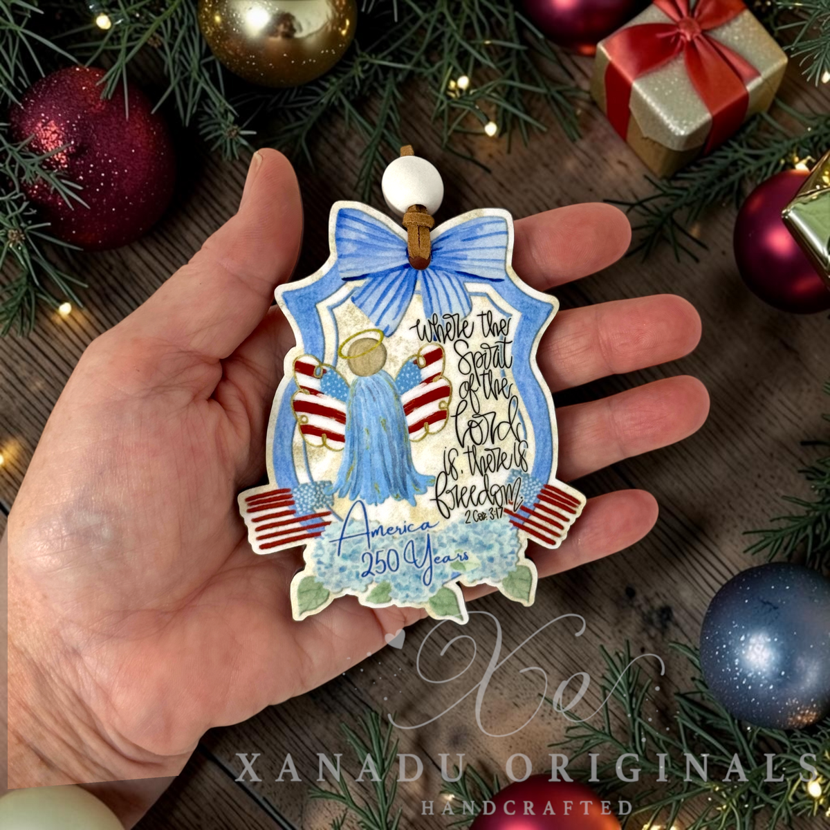 A hand holds a handcrafted Christmas ornament shaped like a bell, decorated with a blue bow, angel figure, red and white stripes, and the text 'America 250 Years' and 'Where the Spirit of the Lord is, there is freedom.' The background shows a rustic wooden surface with pine branches and holiday ornaments.