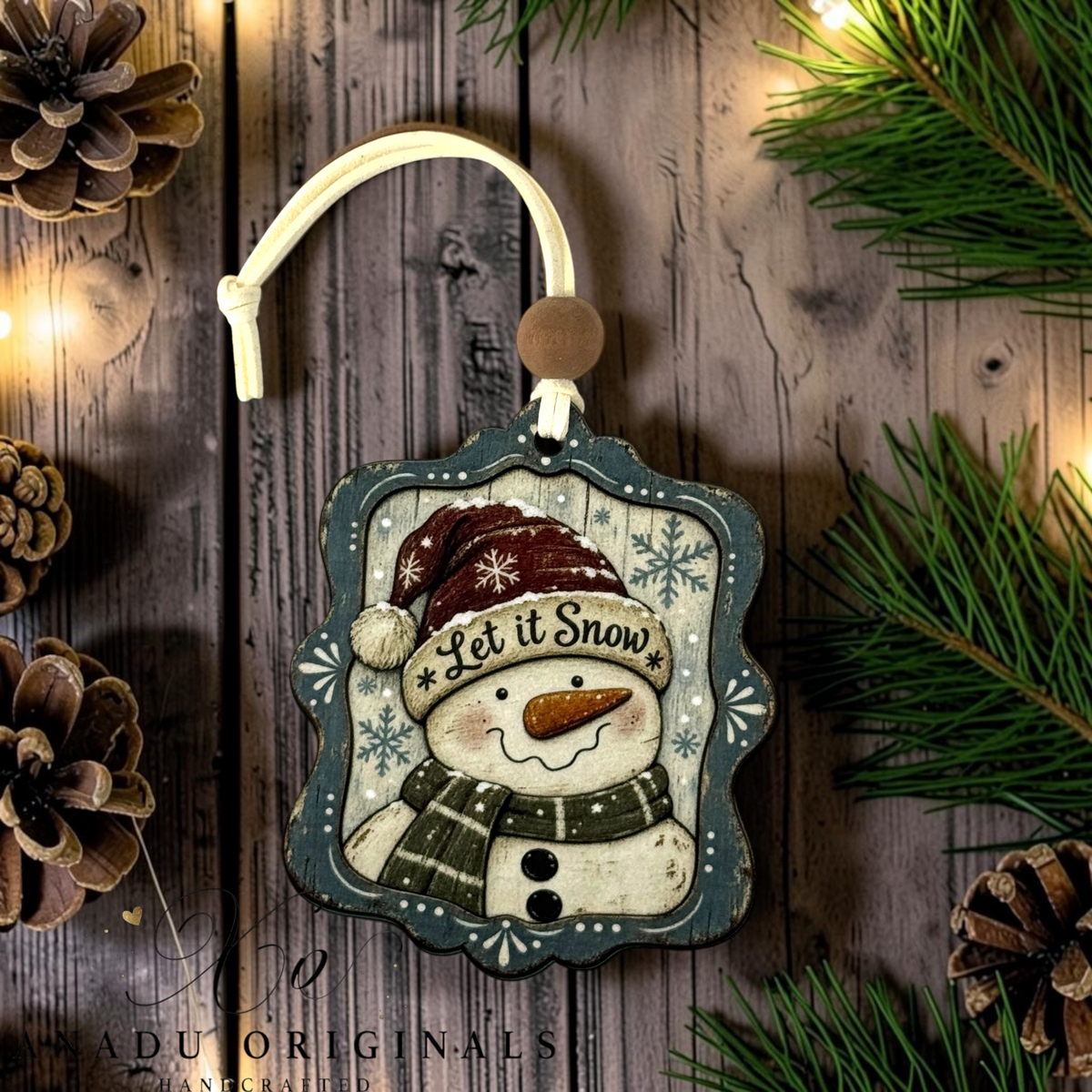 A decorative Christmas ornament featuring a smiling snowman wearing a maroon hat and green scarf, with the words “Let it Snow,” hanging from a cream cord against a rustic wood background with pine branches and pinecones.