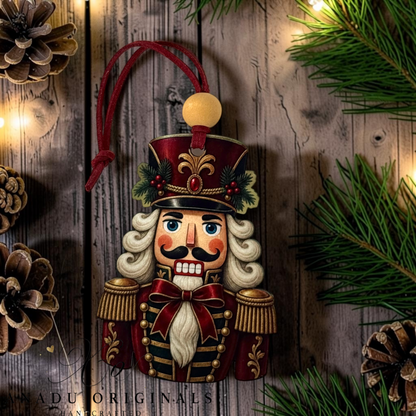 A hand-painted wooden nutcracker Christmas ornament hanging against a rustic wooden background with pine branches, pinecones, and warm string lights. The ornament features a red and gold uniform, white hair and beard, and a red hanging loop.
