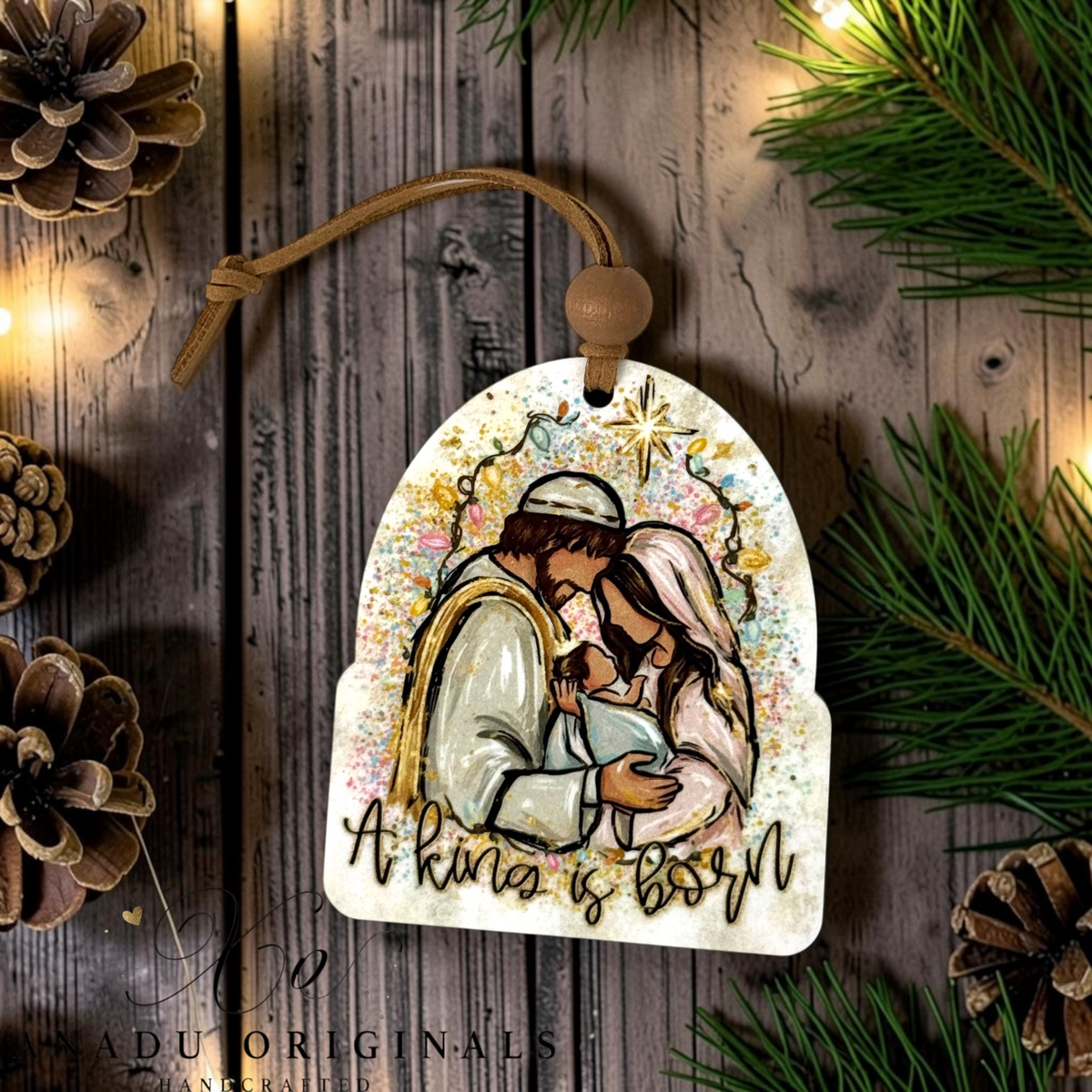 A Christmas ornament featuring an illustration of a baby Jesus in a manger with a star above and the text 'A king is born' at the bottom. The ornament is decorated with colorful paint splatters and hangs from a brown cord, set against a rustic wooden background with pine branches, pinecones, and warm string lights.