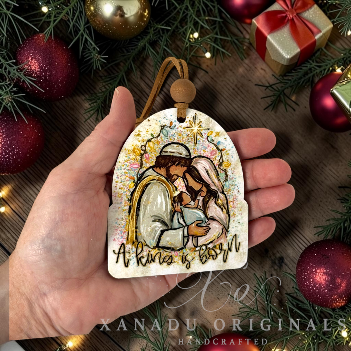 A hand holds a handcrafted Christmas ornament shaped like a wooden tag, featuring an illustration of Jesus and Mary with gold glitter accents and the text 'A King is born.' The background is a rustic wooden surface decorated with red ornaments, green pine branches, and a gold gift box.